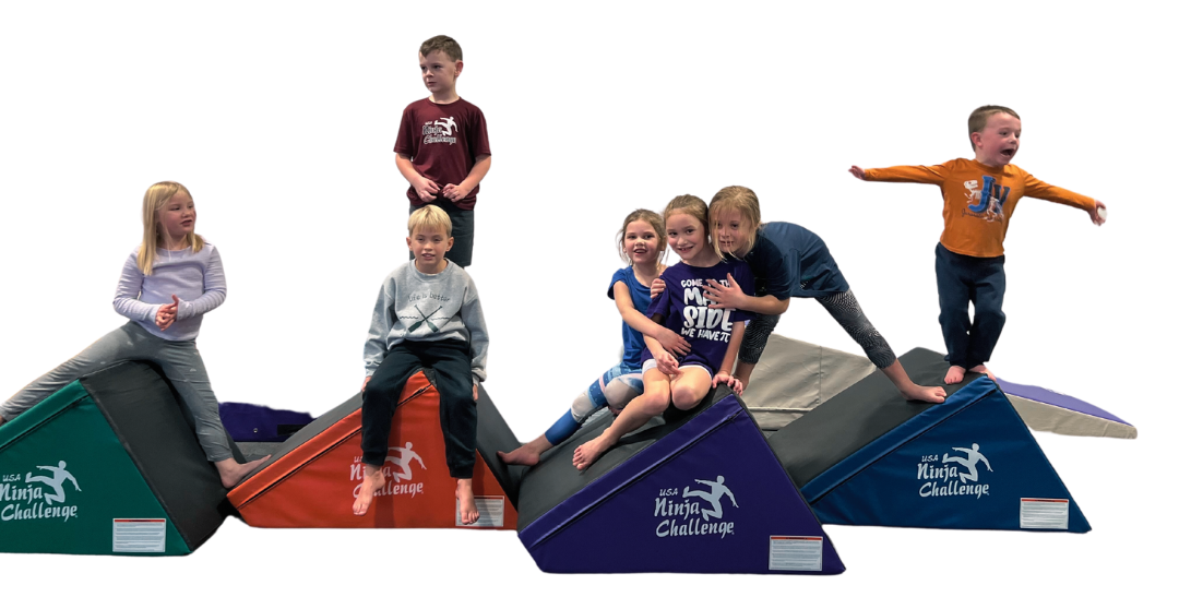 Seven happy children sitting and standing on colorful triangular wedge mats branded with the USA Ninja Challenge logo.