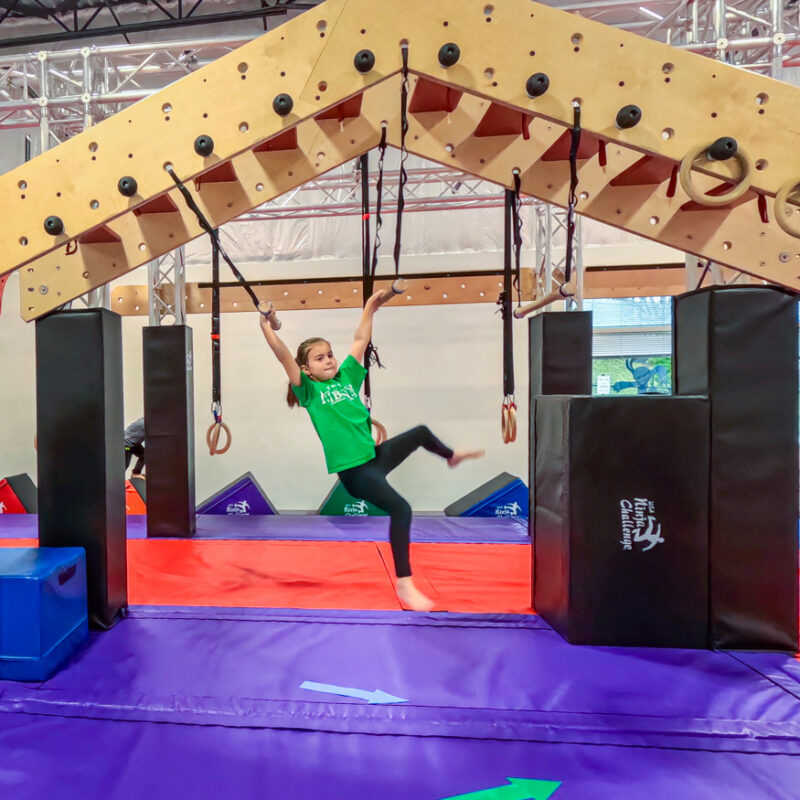 A young girl in a green t-shirt swinging from rings on a wooden ninja obstacle at USA Ninja Challenge