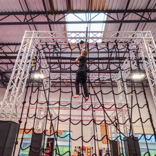 A young boy in black athletic wear climbing a large rope cargo net obstacle towards the ceiling at Ninja Stafford.