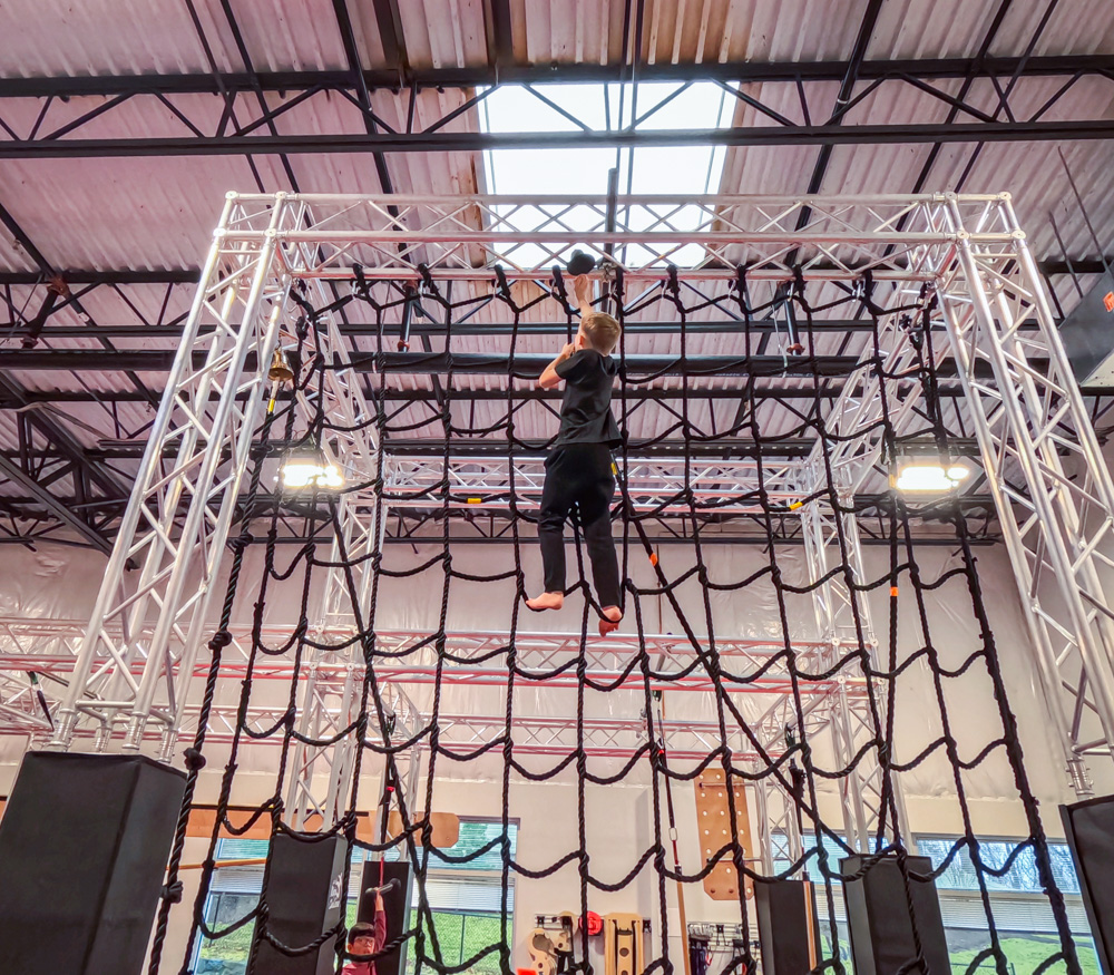 A young boy in black athletic wear climbing a large rope cargo net obstacle towards the ceiling at Ninja Stafford.