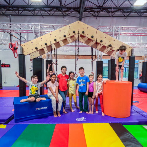 A group of smiling children posing together on colorful gym mats in front of a wooden ninja obstacle at USA Ninja Challenge Stafford